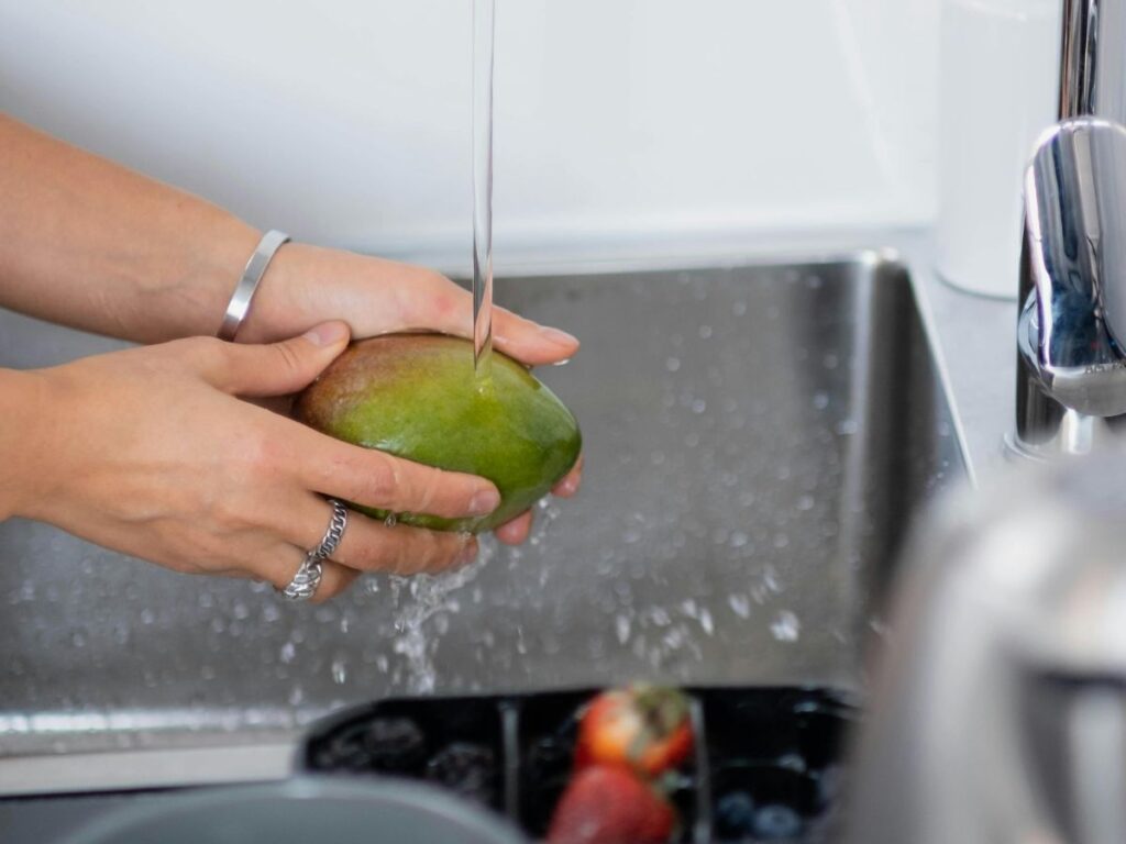 Washing mango at sink – emphasizes clean water for all produce.
